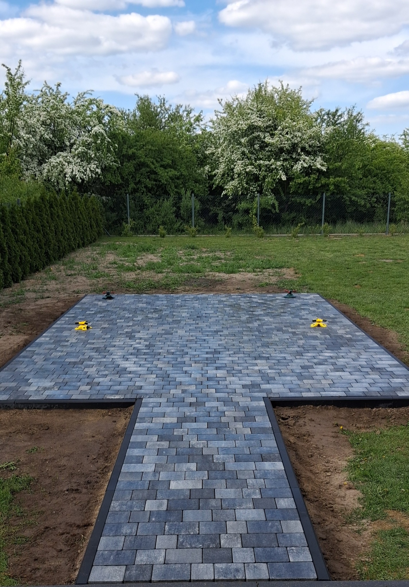 Ułożona kostka brukowa w kształcie litery T na trawniku, z czarnym obramowaniem i żółtymi narzędziami, gotowa do dalszych prac wykończeniowych.