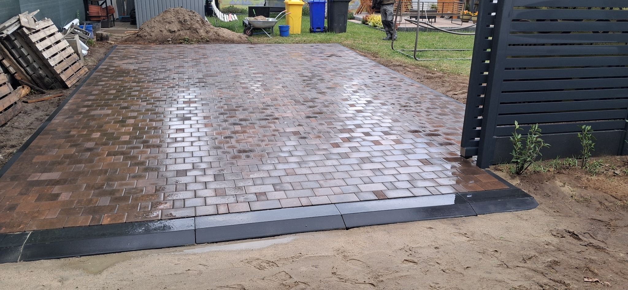 Świeżo ułożona, brązowa kostka brukowa z czarnym obrzeżem, częściowo mokra po deszczu, przylegająca do nowoczesnego, grafitowego ogrodzenia panelowego.