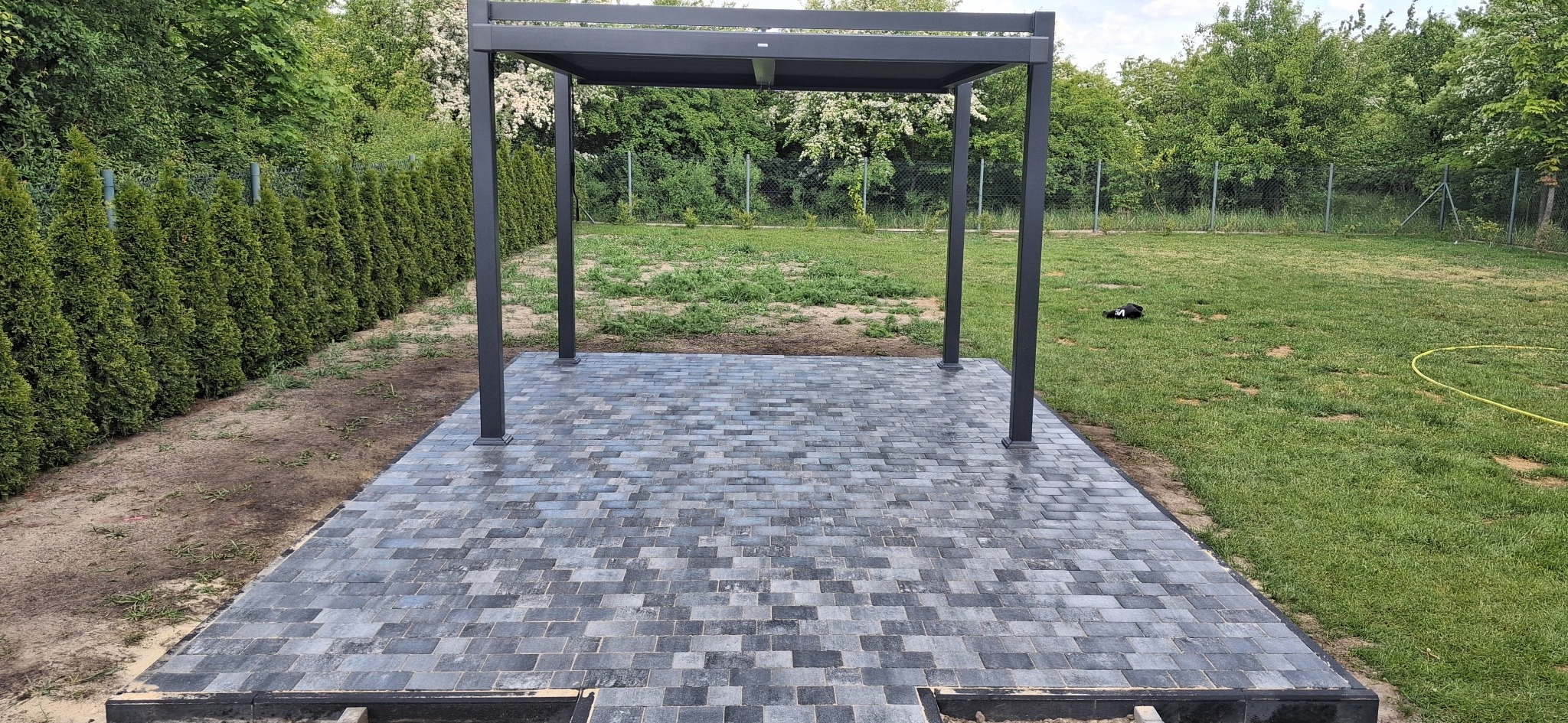Nowoczesna pergola na wybrukowanym tarasie z szaro-niebieskiej kostki, otoczona zielenią. Widoczne thuje i trawnik. Częściowo ukończona realizacja.
