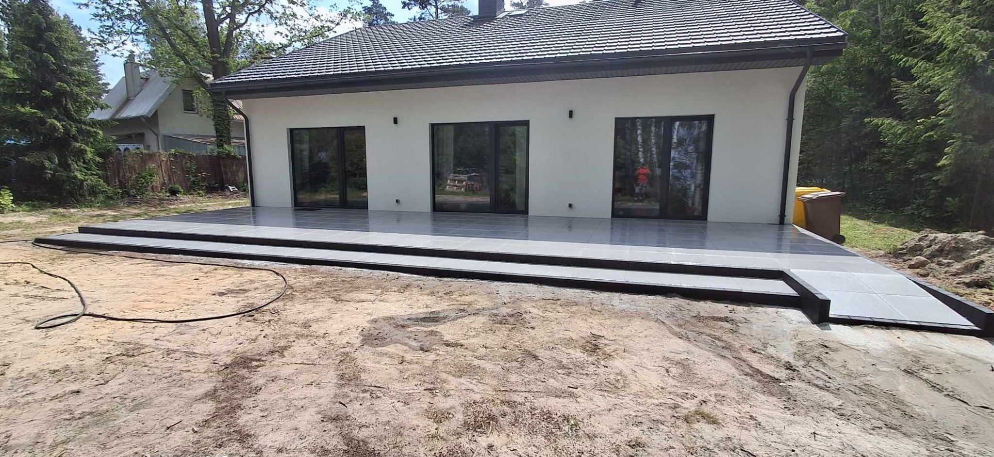 Nowoczesny taras z szarych płyt, z trzema stopniami i rampą, przylegający do białego budynku z trzema oknami. Na pierwszym planie piaszczyste podłoże.