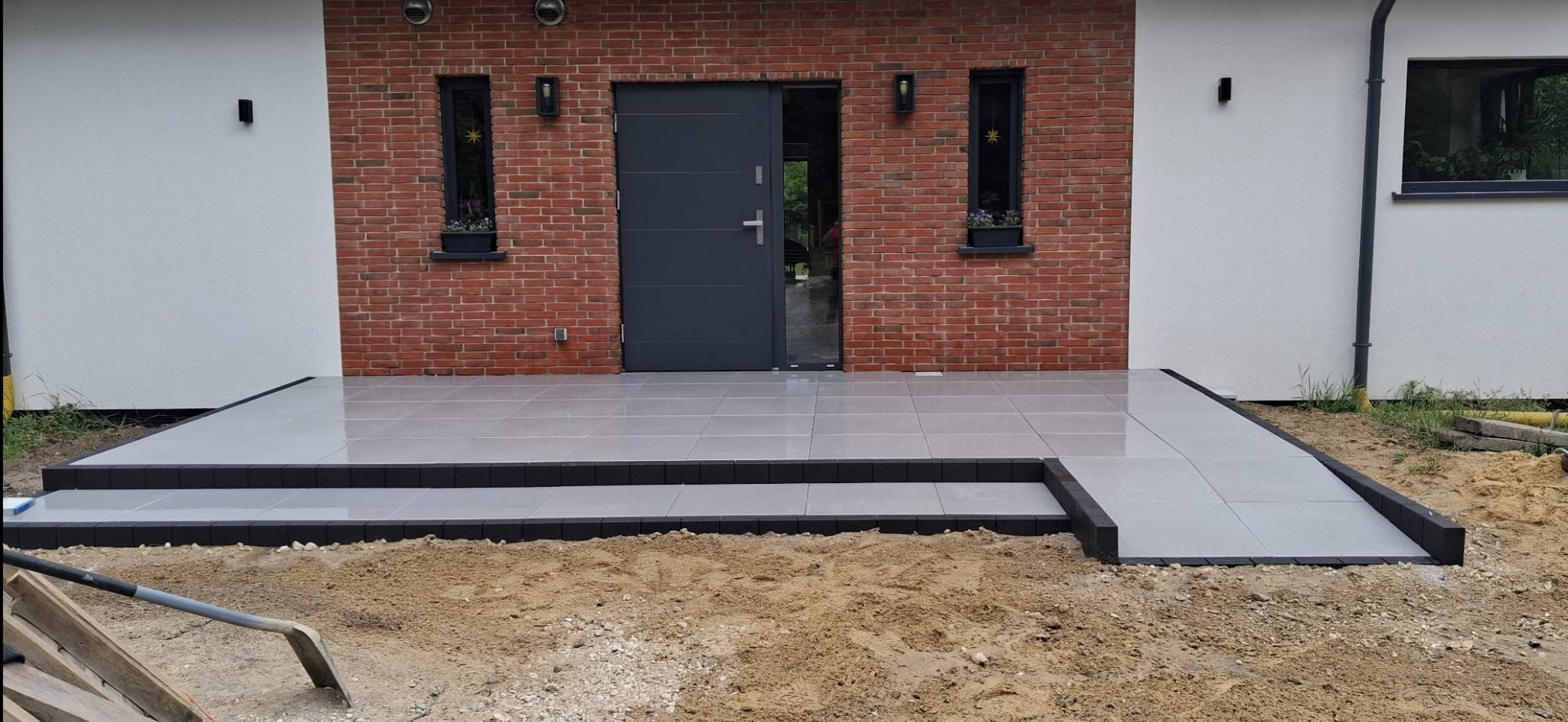 Wykończony taras z szarych płytek ceramicznych przed domem z ceglaną elewacją, widoczne schody i podjazd dla wózków. Na pierwszym planie narzędzie brukarskie.