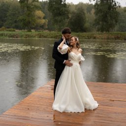 Piotr Krukowski - Para młoda w eleganckich strojach pozuje na drewnianym pomoście nad jeziorem w deszczowy dzień, drzewa w tle.