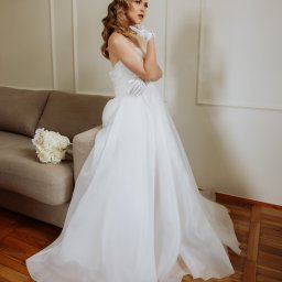 Piotr Krukowski - Panna młoda w białej sukni ślubnej z trenem i rękawiczkach, pozująca obok bukietu i sofy na drewnianej podłodze.