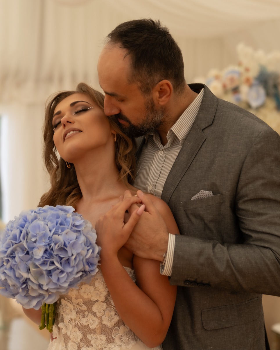 Para młoda: mężczyzna w szarym garniturze obejmuje kobietę w białej sukni z bukietem niebieskich hortensji, wzruszający moment.