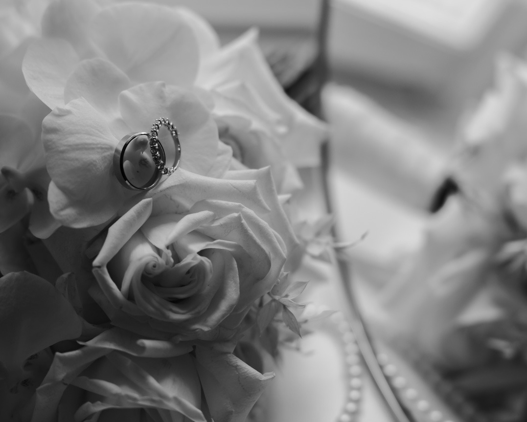 Czarno-białe zbliżenie dwóch obrączek ślubnych spoczywających na białych różach i orchideach, z delikatnym odbiciem perłowego naszyjnika w tle.