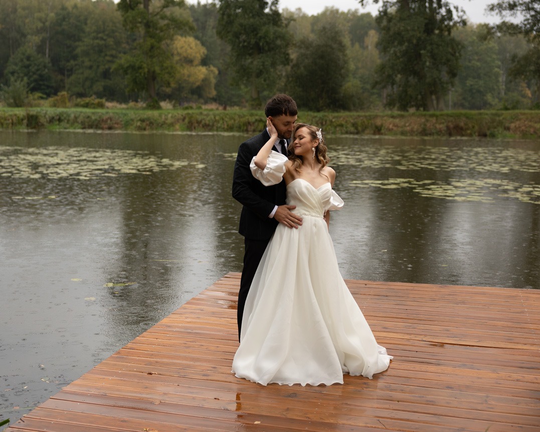 Para młoda w eleganckich strojach pozuje na drewnianym pomoście nad jeziorem w deszczowy dzień, drzewa w tle.