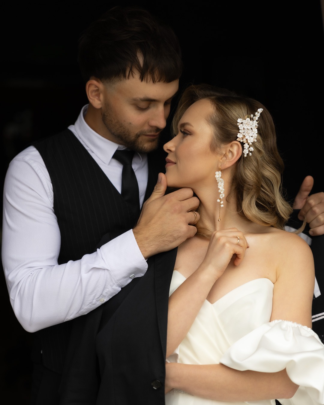 Elegancka para młoda w zbliżeniu, panna młoda z kwiatowym akcentem we włosach i delikatnymi kolczykami, pan młody w klasycznym garniturze, romantyczne spojrzenie.