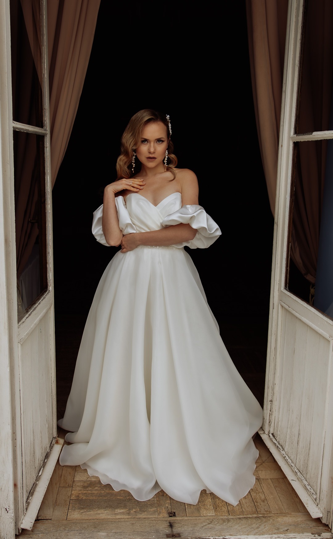 Panna młoda w białej sukni z odkrytymi ramionami i bufkami, pozuje w otwartych, białych drzwiach z beżowymi zasłonami w tle.