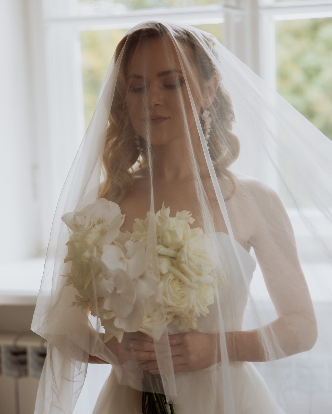 Panna młoda z zamkniętymi oczami, częściowo zasłonięta welonem, trzymająca bukiet białych róż i orchidei na tle okna.
