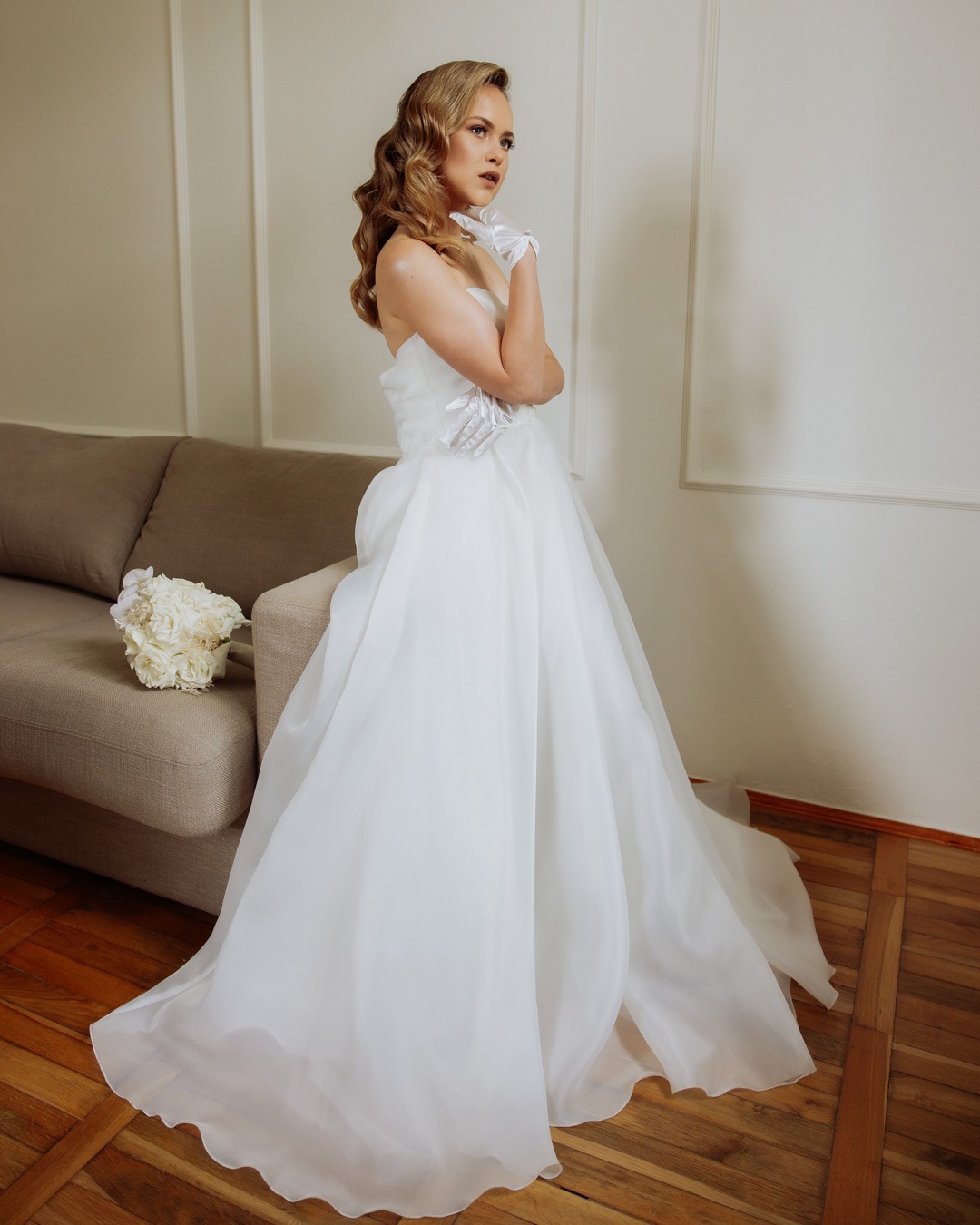 Panna młoda w białej sukni ślubnej z trenem i rękawiczkach, pozująca obok bukietu i sofy na drewnianej podłodze.