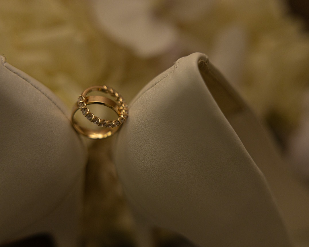 Złoty pierścionek z brylantami leżący na kremowych, skórzanych szpilkach ślubnych na tle bukietu z jasnych kwiatów.