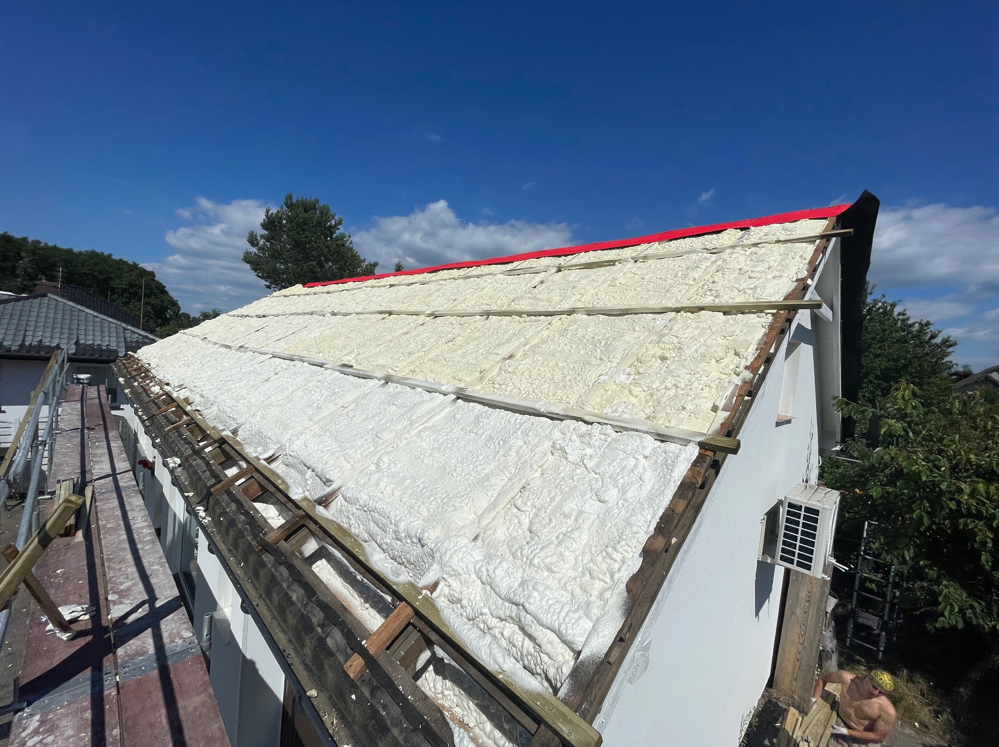 Poddasze domu jednorodzinnego z widoczną warstwą izolacji pianką PUR, drewniane łaty na dachu, czerwony pas zabezpieczający, błękitne niebo w tle.