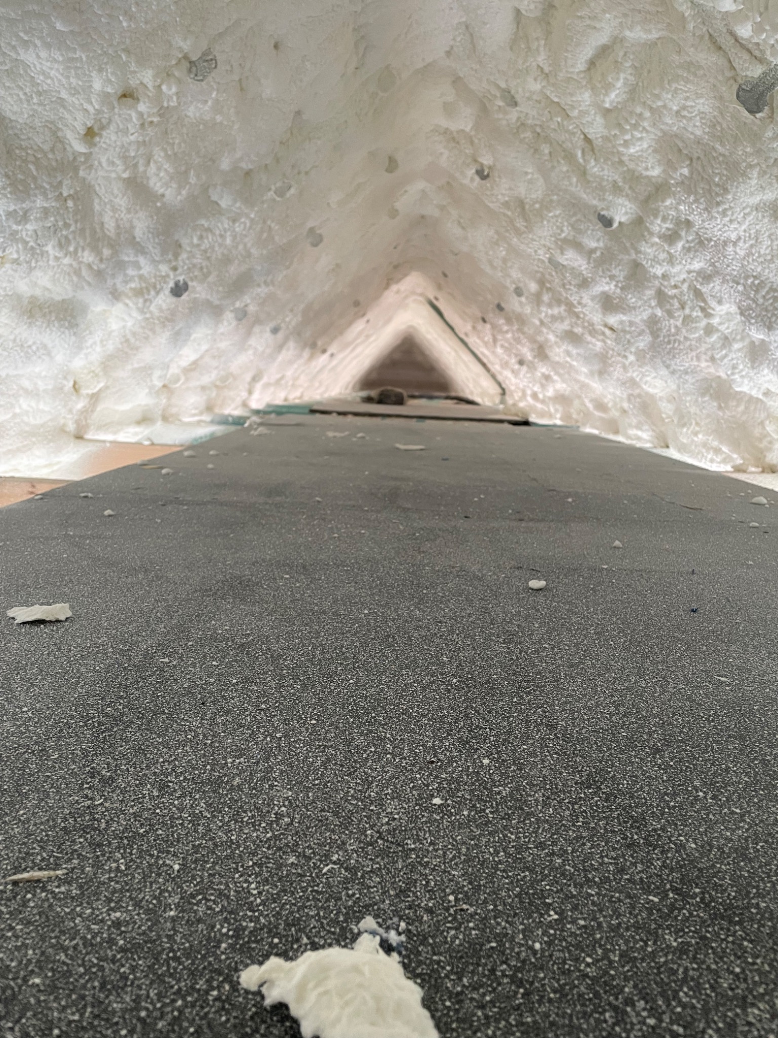 Poddasze z widoczną szarą powierzchnią i nałożoną białą, gęstą pianą izolacyjną na skośnych ścianach, tworzącą trójkątny tunel.