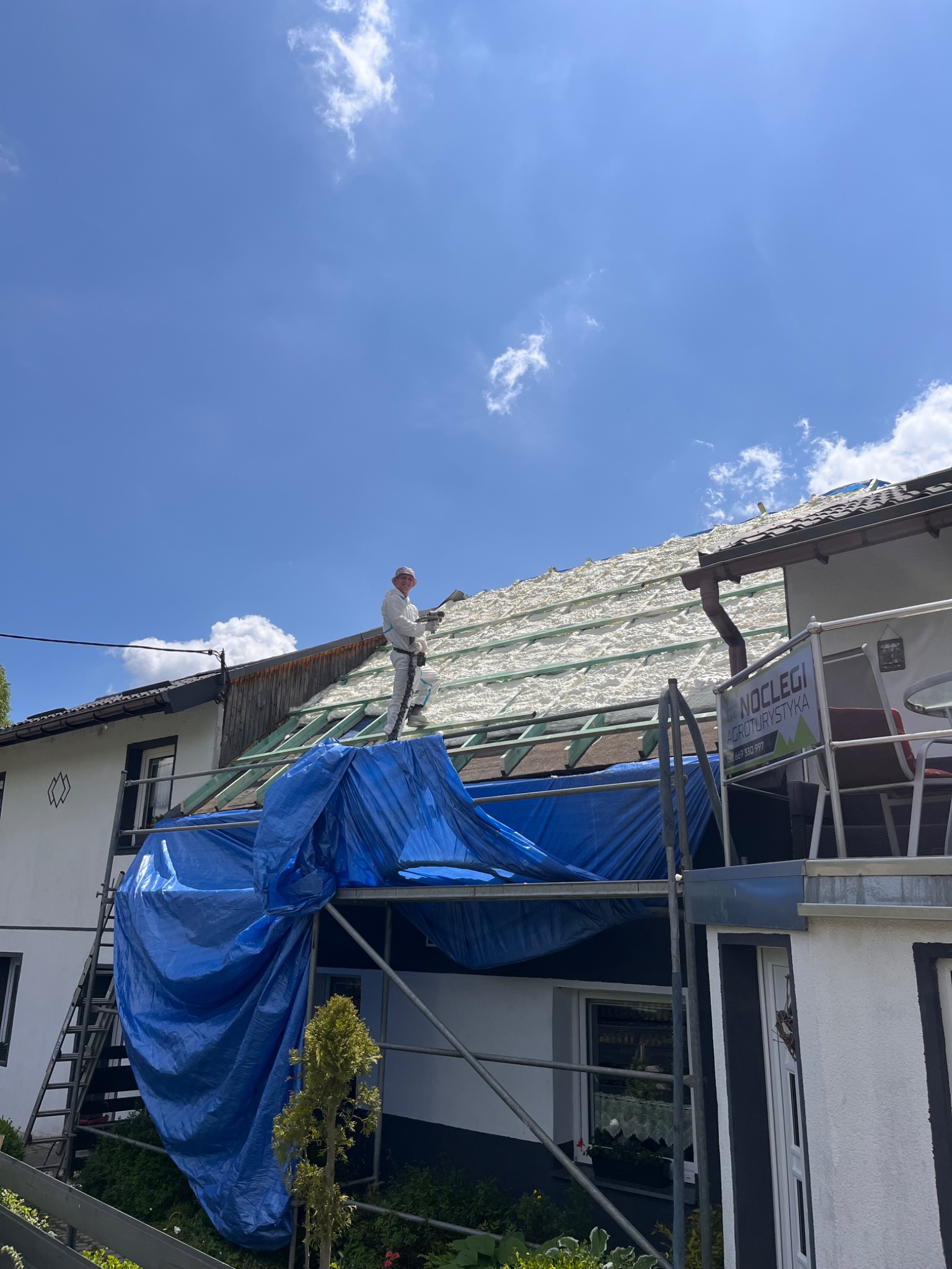 Pracownik w kombinezonie ochronnym aplikuje piankę izolacyjną na drewnianą konstrukcję dachu domu jednorodzinnego, widoczne rusztowanie, błękitne niebo z chmurami w tle.