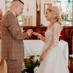 Para młoda podczas ceremonii ślubnej w kościele, wymiana obrączek, on w brązowym garniturze w kratę, ona w białej sukni z koronkowymi rękawami, witraż w tle.