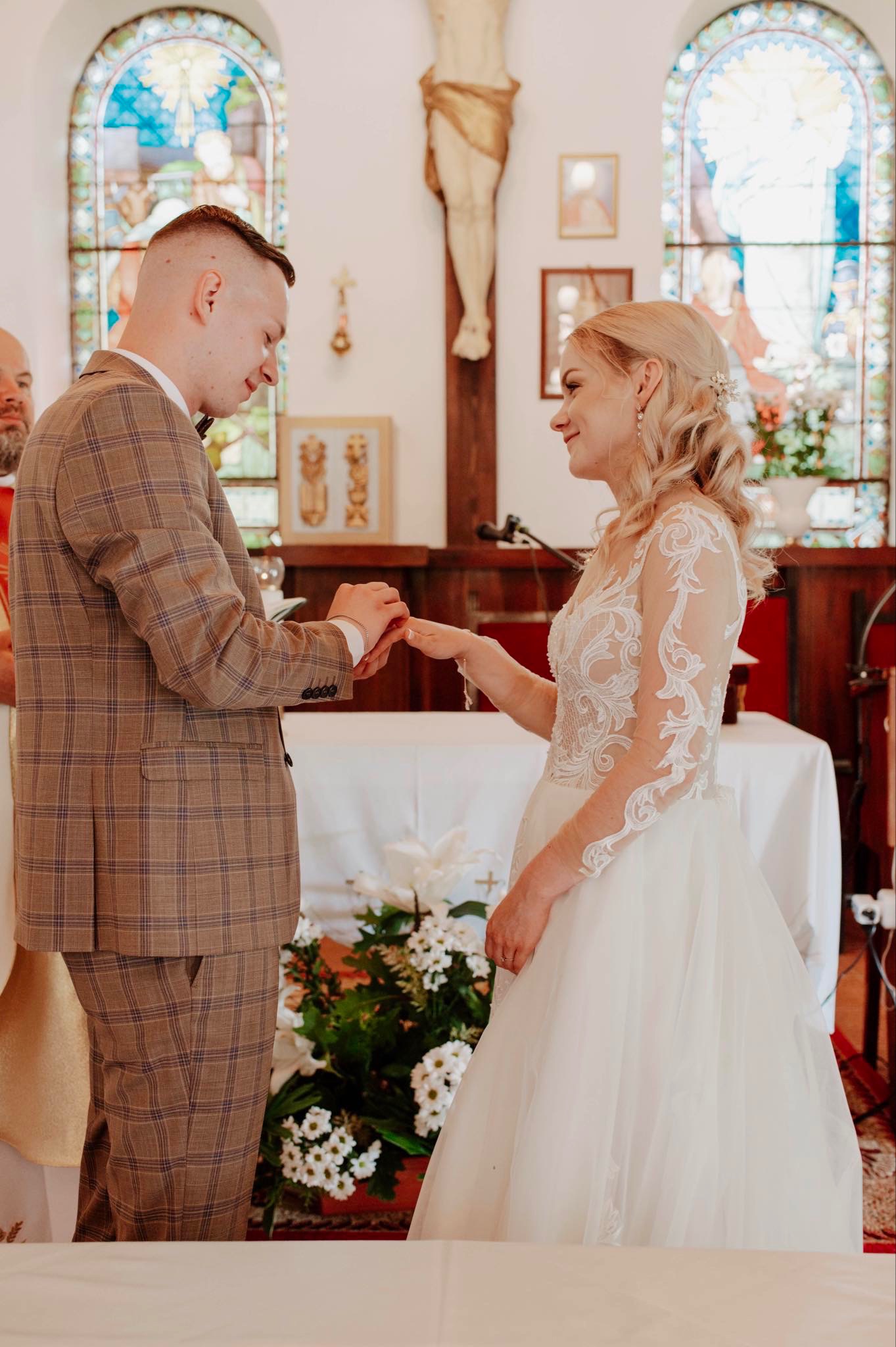 Para młoda podczas ceremonii ślubnej w kościele, wymiana obrączek, on w brązowym garniturze w kratę, ona w białej sukni z koronkowymi rękawami, witraż w tle.