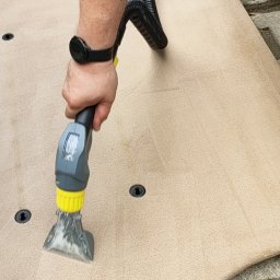 Ivan Sosiedko - Czyszczenie beżowego dywanu odkurzaczem piorącym z żółtą końcówką, widoczna woda i czyszczona powierzchnia, na betonowym podłożu.