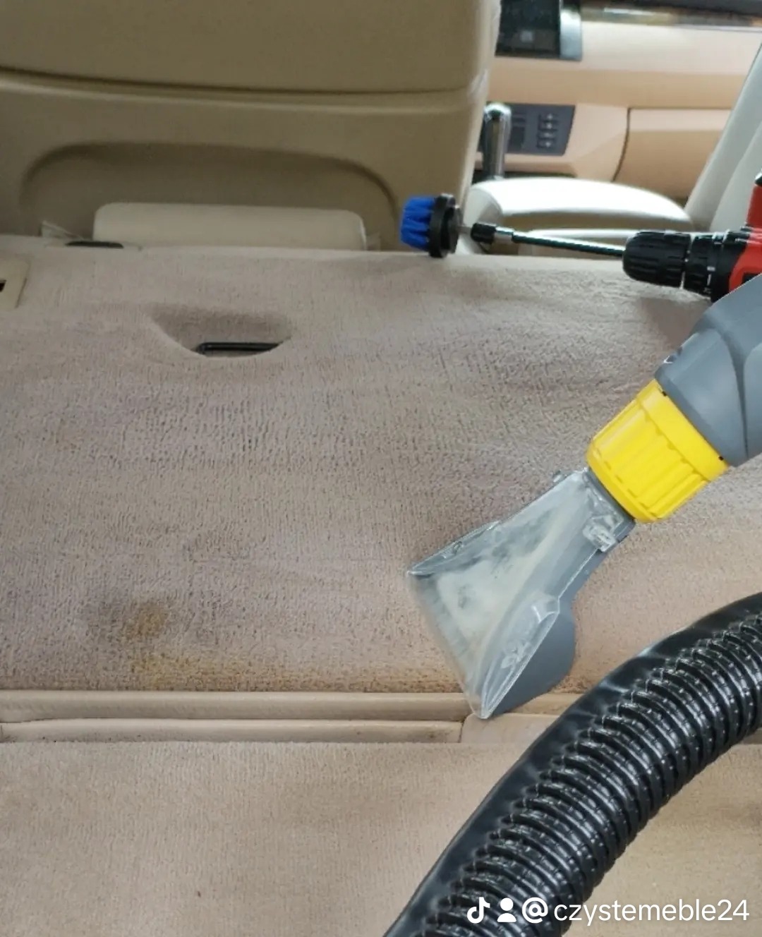 Czyszczenie beżowej tapicerki samochodowej odkurzaczem piorącym z żółtą końcówką, widoczne zabrudzenia na materiale i wąż odkurzacza.