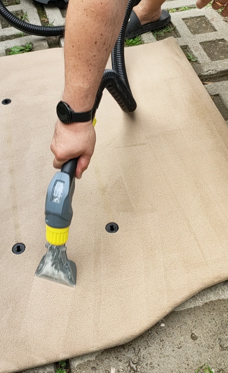Czyszczenie beżowego dywanu odkurzaczem piorącym z żółtą końcówką, widoczna woda i czyszczona powierzchnia, na betonowym podłożu.