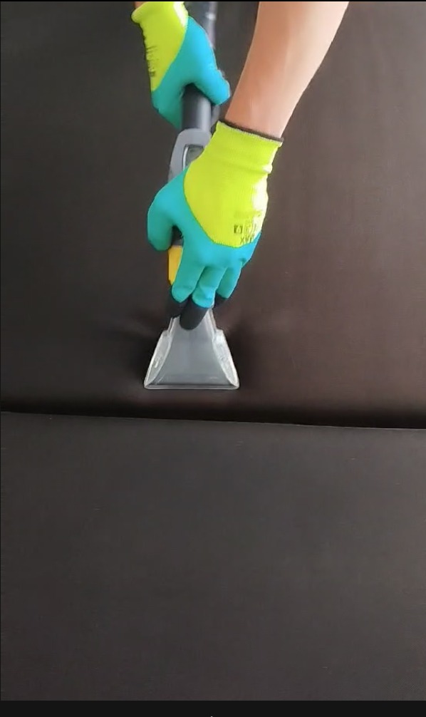 Czyszczenie ciemnobrązowej sofy za pomocą odkurzacza z przezroczystą końcówką i rąk w zielono-niebieskich rękawicach ochronnych.