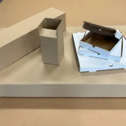 Różnorodne, rozłożone kartonowe pudełka: długie prostokątne, kwadratowe i białe pudełka na pizzę, ułożone na dużym arkuszu tektury falistej.