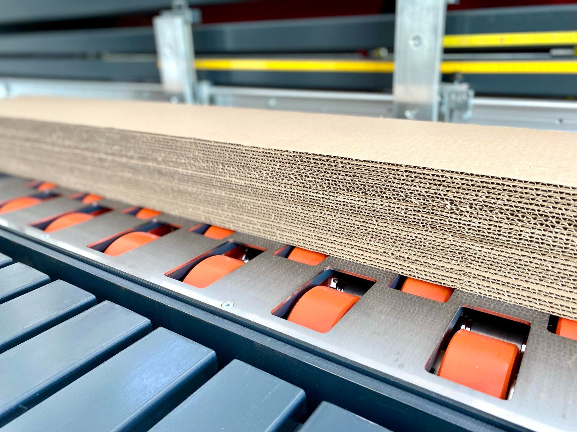 Stos tektury falistej na linii produkcyjnej z pomarańczowymi rolkami podajnika, widok z bliska na strukturę tektury.