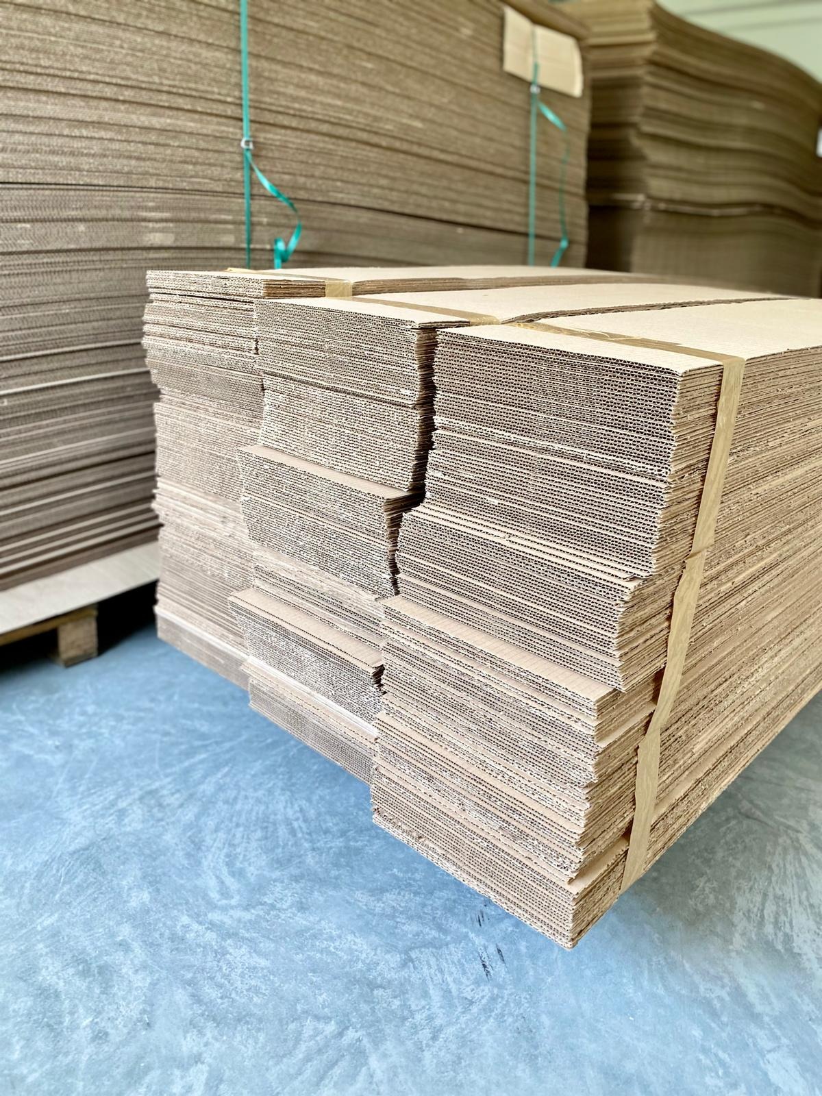 Stosy płaskich, rozłożonych kartonów na paletach, zabezpieczone taśmą, gotowe do użycia w magazynie.