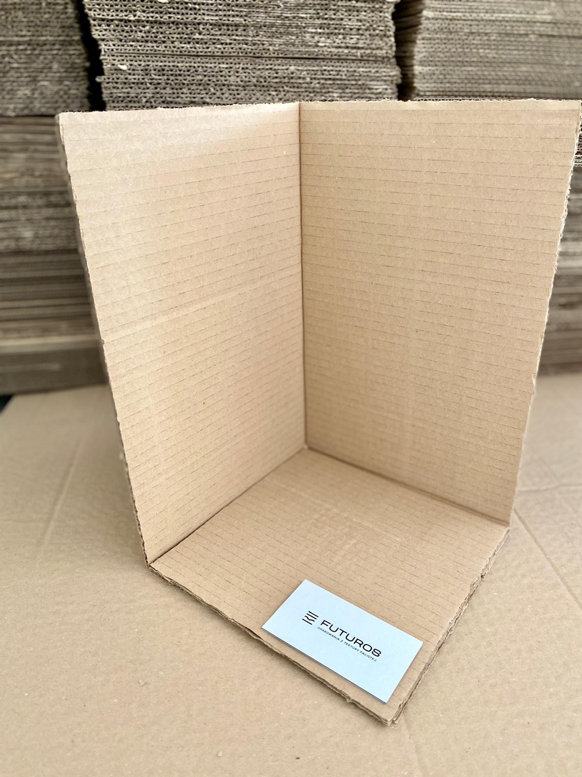 Rozłożony kartonowy narożnik z widoczną wizytówką firmy Futuros na tle stosu arkuszy tektury falistej.