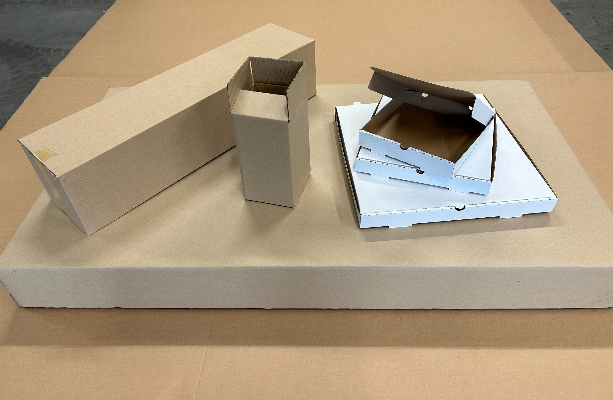 Różnorodne, rozłożone kartonowe pudełka: długie prostokątne, kwadratowe i białe pudełka na pizzę, ułożone na dużym arkuszu tektury falistej.