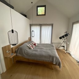 FIRMA USŁUGOWO-REMONTOWA Bartłomiej Sprawka - Jasna sypialnia na poddaszu z drewnianą podłogą, łóżkiem z szarą narzutą i poduszkami z motywem jelenia, nowoczesnymi lampami wiszącymi w kształcie klatek oraz teleskopem na statywie przy oknie.