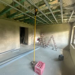 FIRMA USŁUGOWO-REMONTOWA Bartłomiej Sprawka - Pomieszczenie w trakcie remontu z zamontowanym metalowym stelażem sufitu podwieszanego, widoczną izolacją natryskową i otynkowanymi ścianami. Na podłodze profile i poziomica laserowa.