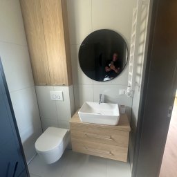 FIRMA USŁUGOWO-REMONTOWA Bartłomiej Sprawka - Nowoczesna toaleta z podwieszanym WC, drewnianą szafką z umywalką i okrągłym lustrem. Białe płytki ścienne i podłogowe, obok wysoka szafka w kolorze drewna. W lustrze odbicie osoby robiącej zdjęcie.