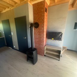FIRMA USŁUGOWO-REMONTOWA Bartłomiej Sprawka - Nowoczesne wnętrze z czarnym piecem wolnostojącym, ceglanym kominem, ścianą z efektem betonu, drewnianymi belkami sufitowymi i jasnymi płytkami imitującymi drewno. Telewizor w folii, ciemne drzwi.