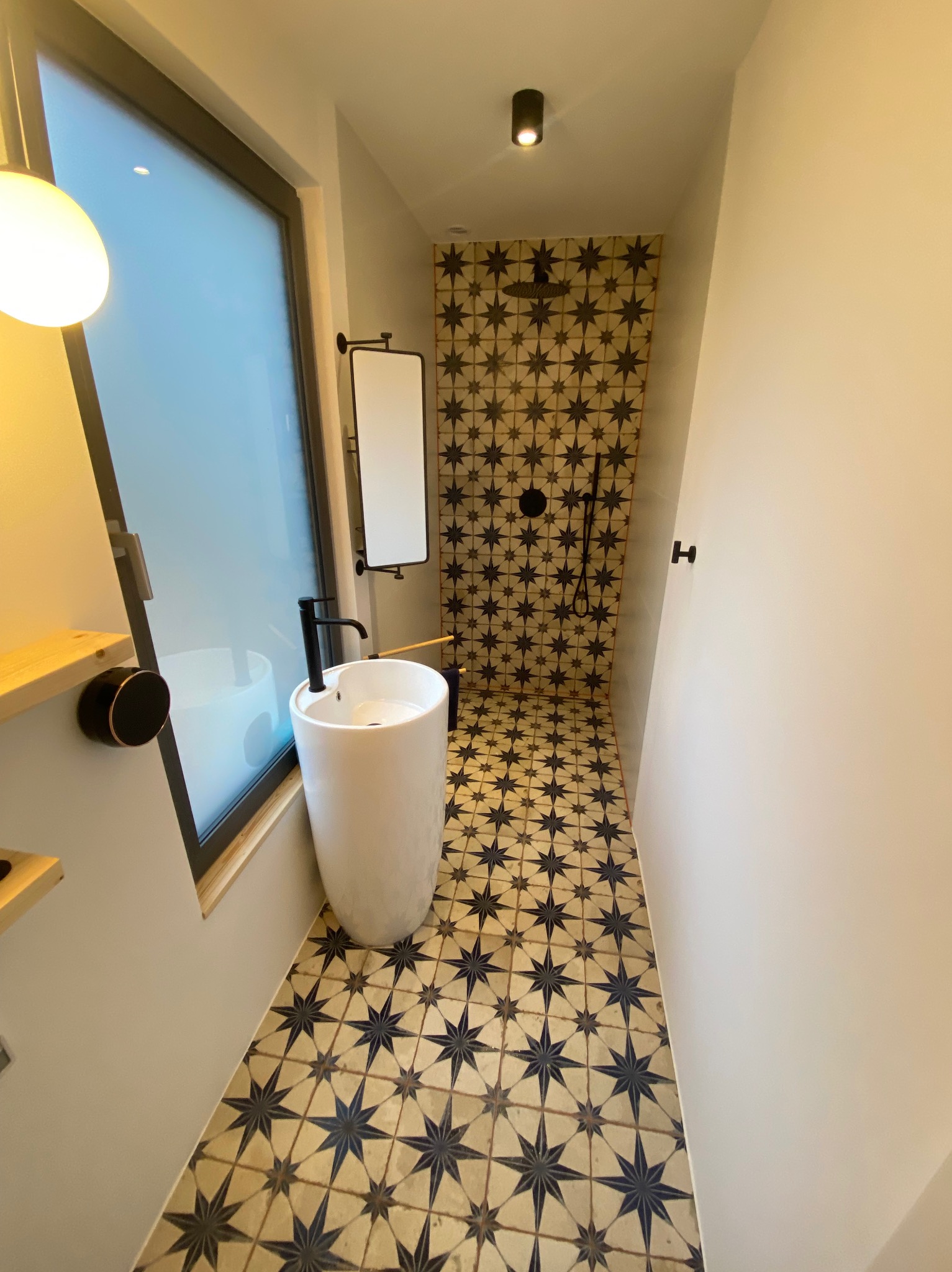 Wąska łazienka z kabiną prysznicową wykończona płytkami w gwiazdy, biała umywalka nablatowa na postumencie i czarna armatura.