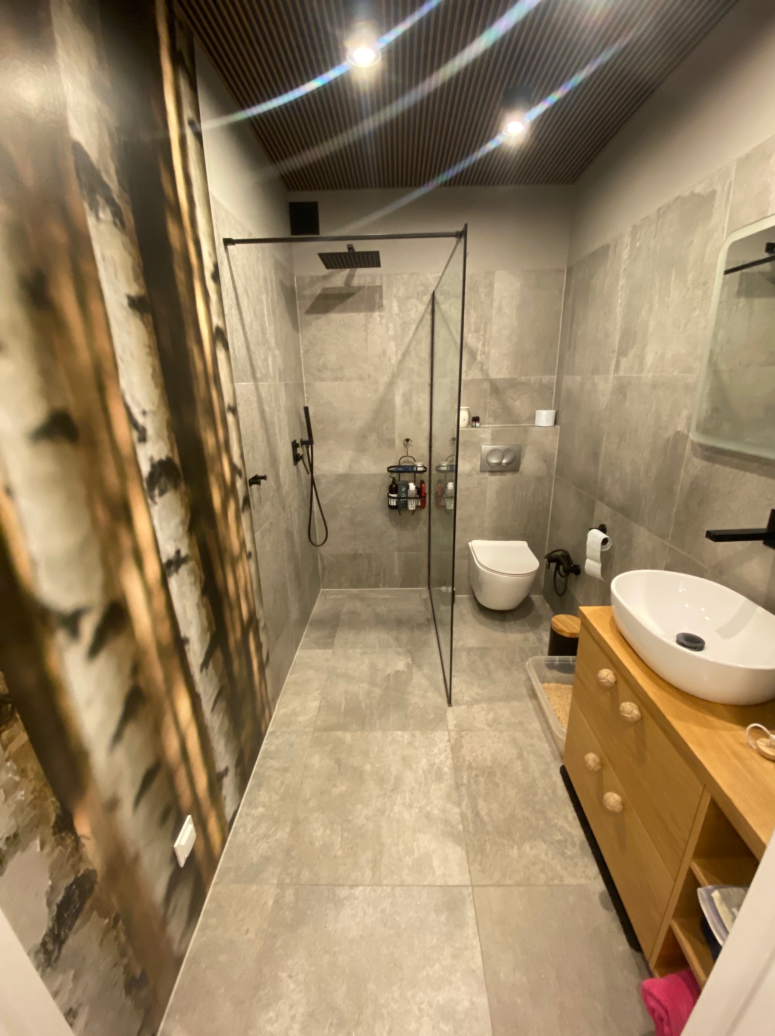 Nowoczesna łazienka z motywem brzozowego lasu na ścianie, szklana kabina prysznicowa z czarnymi elementami, umywalka nablatowa na drewnianej szafce z okrągłymi uchwytami, szare płytki podłogowe...