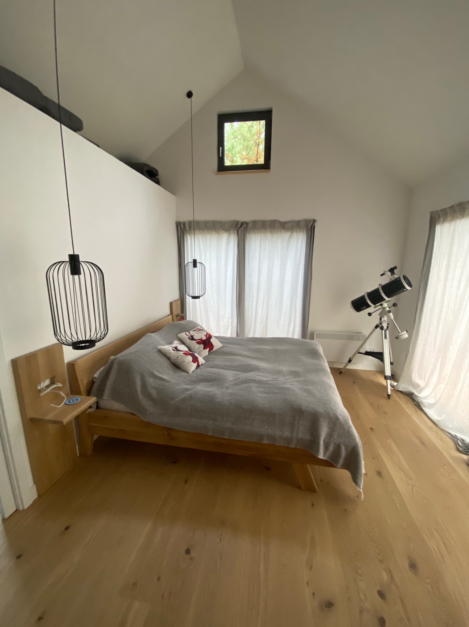 Jasna sypialnia na poddaszu z drewnianą podłogą, łóżkiem z szarą narzutą i poduszkami z motywem jelenia, nowoczesnymi lampami wiszącymi w kształcie klatek oraz teleskopem na statywie przy oknie.