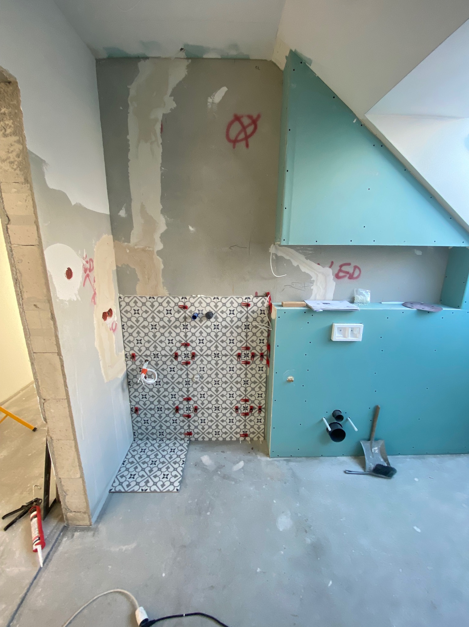 Montaż płytek wzorzystych na ścianie łazienki, widoczne krzyżyki dystansowe, fragment ściany z płyt karton-gips, instalacja sanitarna w tle, surowy stan wykończenia.