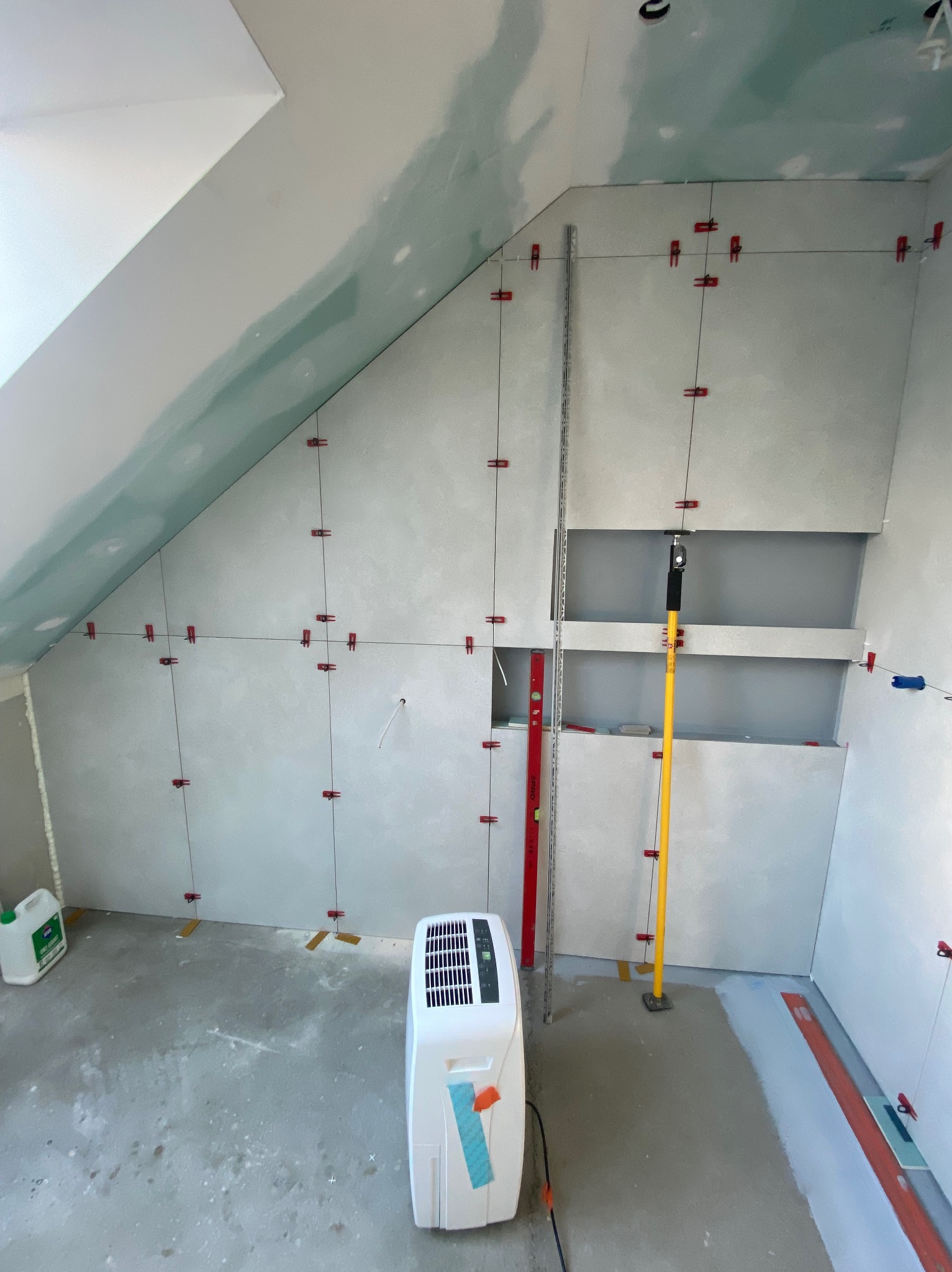 Skośne poddasze w trakcie montażu płyt kartonowo-gipsowych, widoczne profile, poziomice, sznurki traserskie i czerwone klipsy do mocowania, klimatyzator przenośny na podłodze.
