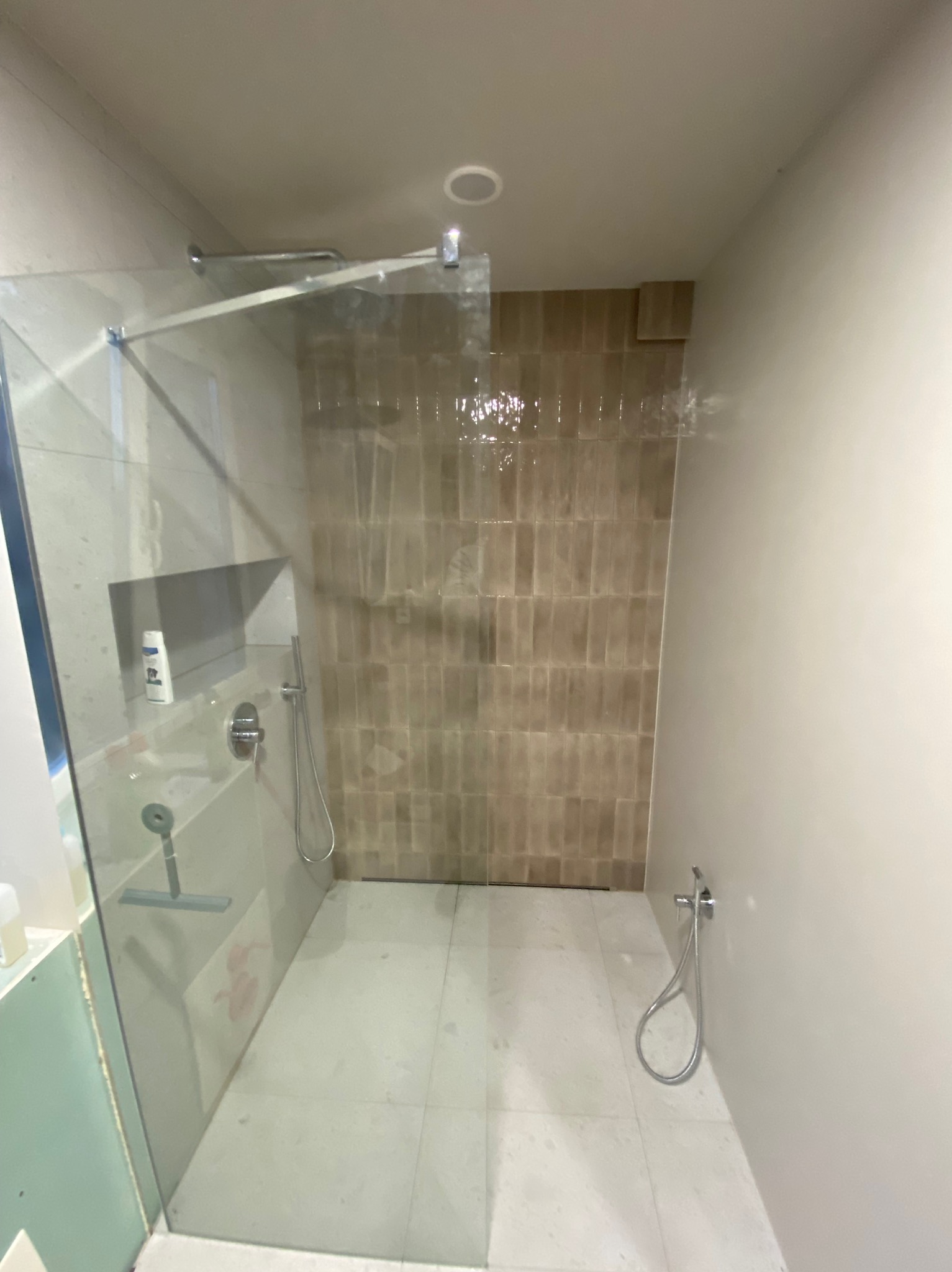 Nowoczesna kabina prysznicowa z beżowymi, pionowo ułożonymi płytkami na ścianie, widoczna półka w ścianie z kosmetykami oraz szklana ściana kabiny.