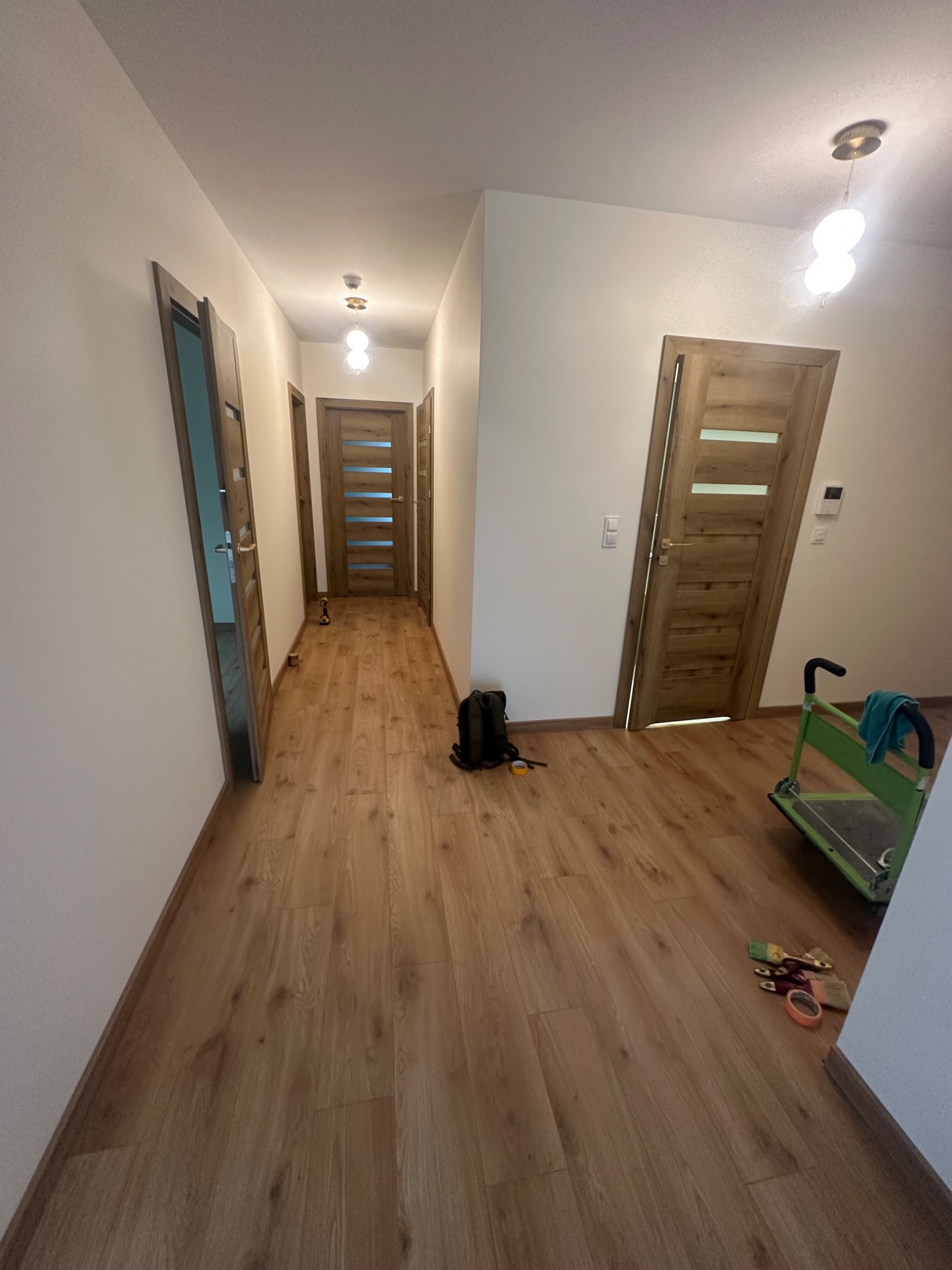 Korytarz po remoncie z drewnianą podłogą i drzwiami. Widoczny wózek transportowy i pędzle malarskie. Oświetlenie sufitowe. Perspektywa z głębi korytarza.
