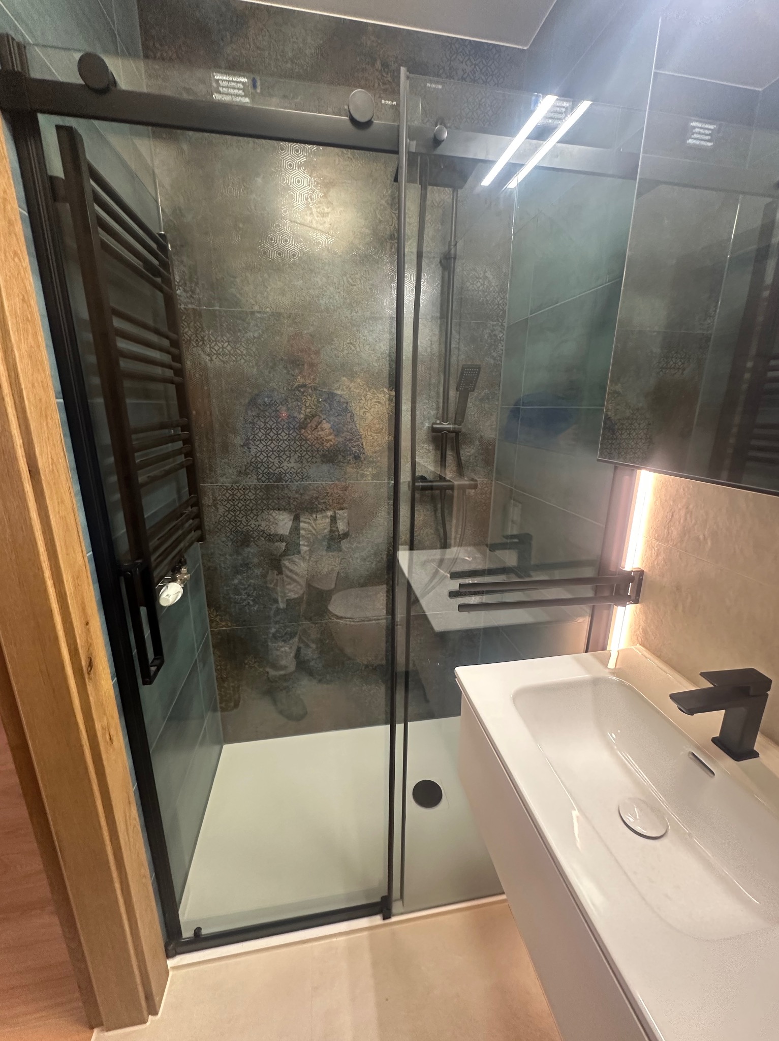 Nowoczesna łazienka po remoncie: kabina prysznicowa z czarnymi ramami, umywalka z szafką, grzejnik drabinkowy. Widoczne płytki w odcieniach szarości i beżu.