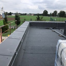 Total Service Usługi Elektryczne - Szary dach płaski z zamontowanymi elementami instalacji odgromowej, widok na okoliczne pola i budynki w pochmurny dzień.