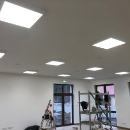 Total Service Usługi Elektryczne - Montaż paneli LED w jasnym pomieszczeniu z białymi ścianami, widoczne drabiny i rusztowania, okna z brązowymi ramami, częściowo widoczna osoba w prawym dolnym rogu.