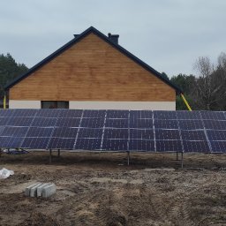 Total Service Usługi Elektryczne - Montaż paneli fotowoltaicznych na gruncie przed domem jednorodzinnym, widoczny pracownik w odzieży roboczej i narzędzia budowlane, w tle las i budynki.