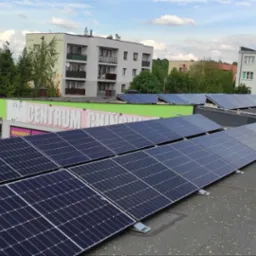 Rząd paneli fotowoltaicznych na dachu budynku handlowego z zielonym dachem, widok z góry na tle zabudowy miejskiej i błękitnego nieba z chmurami.