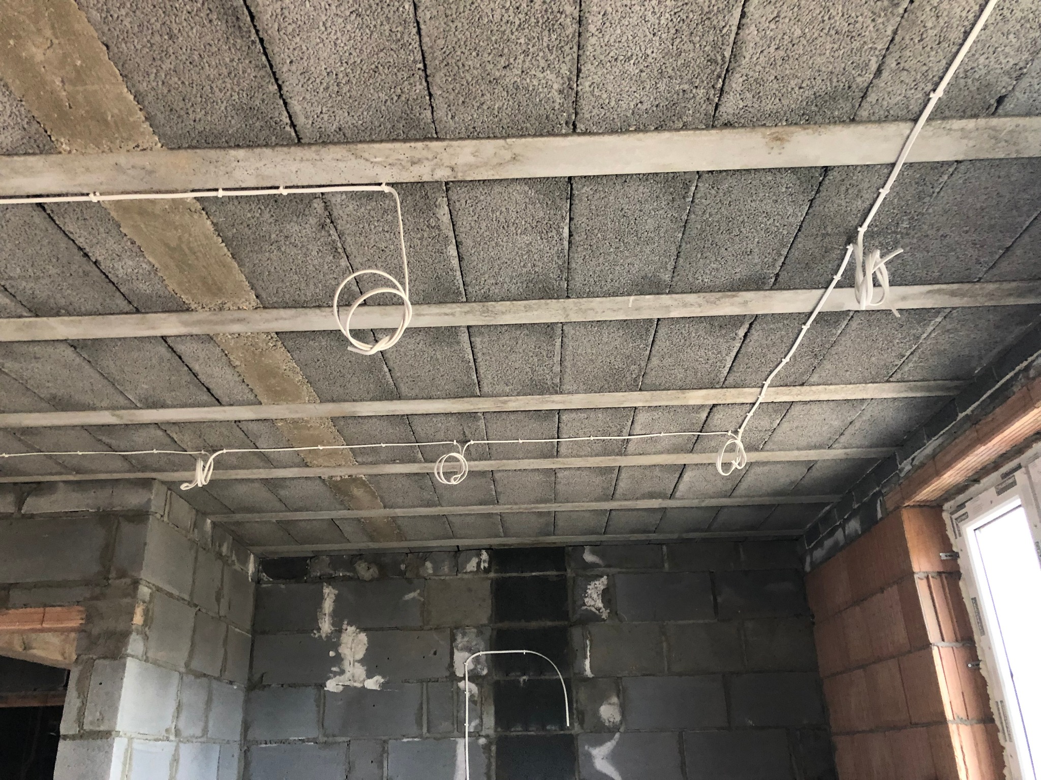Surowy stan wnętrza w trakcie prac wykończeniowych: widoczne przewody elektryczne zamontowane pod betonowym sufitem, ściany z pustaków, okno w narożniku.