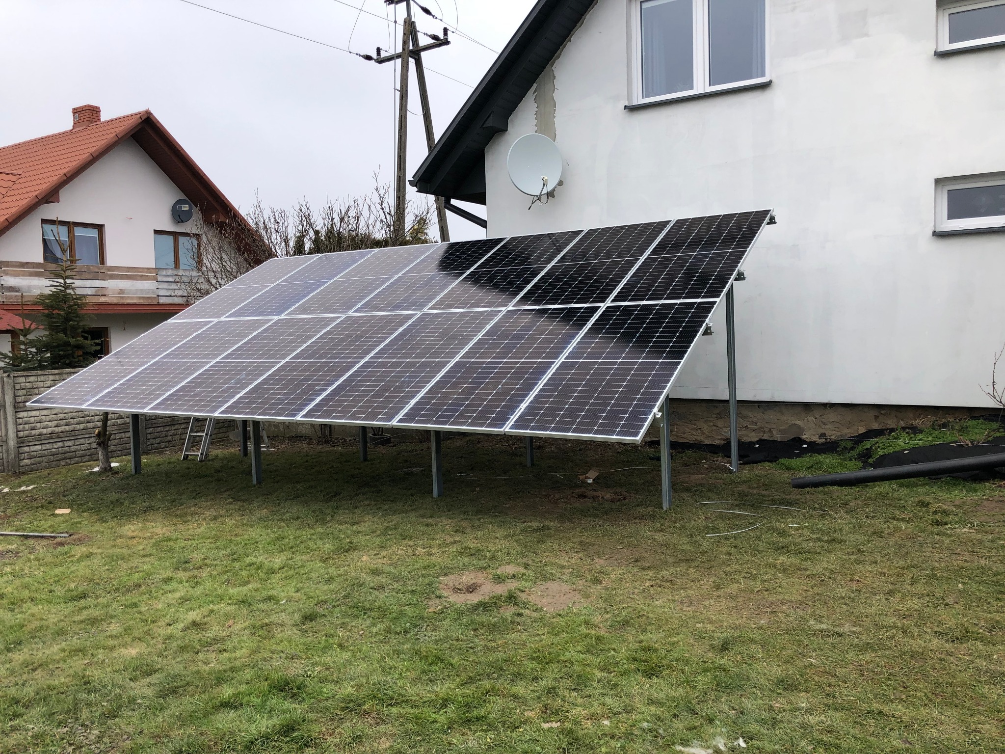 Panel fotowoltaiczny zamontowany na trawniku przed domem z białą elewacją i anteną satelitarną, widoczne słupy i przewody elektryczne.