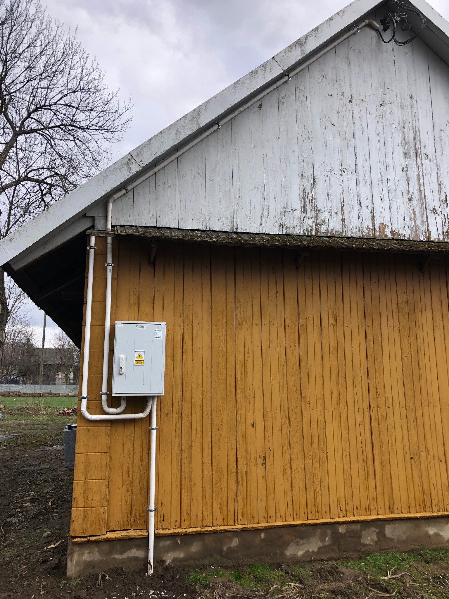 Szara skrzynka elektryczna z symbolem ostrzegawczym wysokiego napięcia, zamontowana na żółtej, drewnianej ścianie budynku z widocznymi rurami PCV i elementami instalacji elektrycznej na zewnątrz.