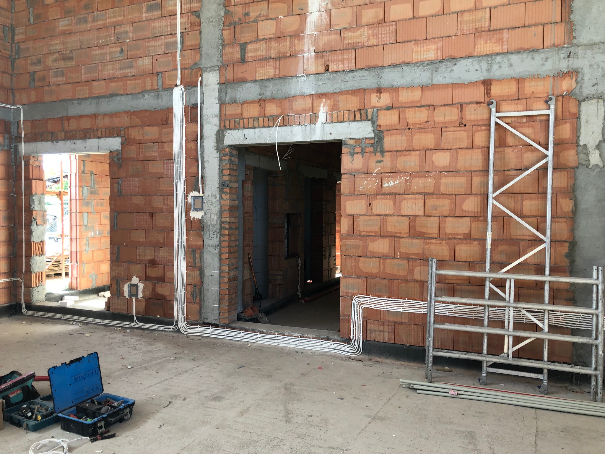 Wewnętrzne ściany z czerwonej cegły z widocznymi, białymi rurami instalacji elektrycznej, otwarte skrzynki z narzędziami i metalowe rusztowanie w surowym stanie wykończenia budynku.