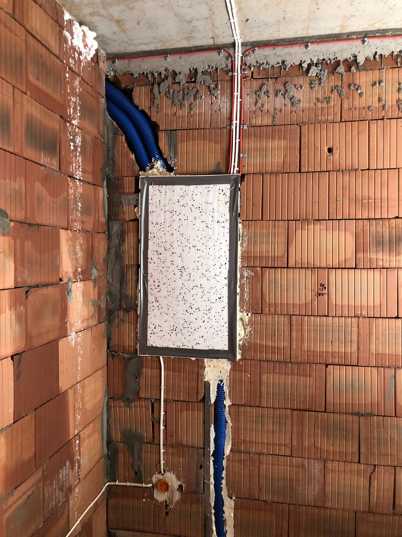 Niedokończona instalacja elektryczna w surowym murze z cegły: białe kable elektryczne, niebieskie rury karbowane, puszka podtynkowa i otwór na rozdzielnicę zabezpieczony kartonem.